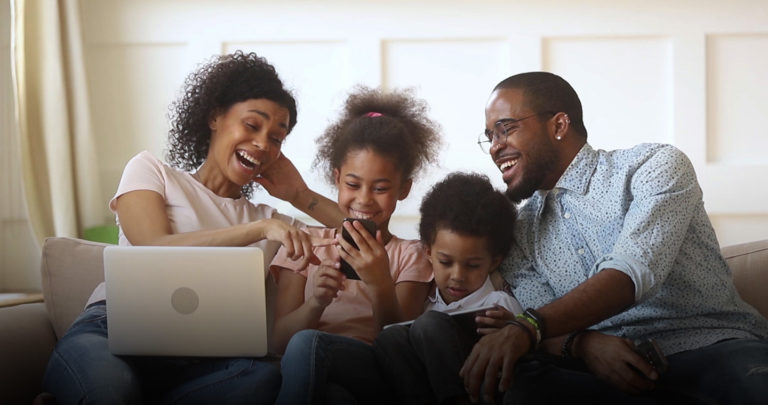 family laughing together on a couch using different type of technology such as phones, tablets and laptops